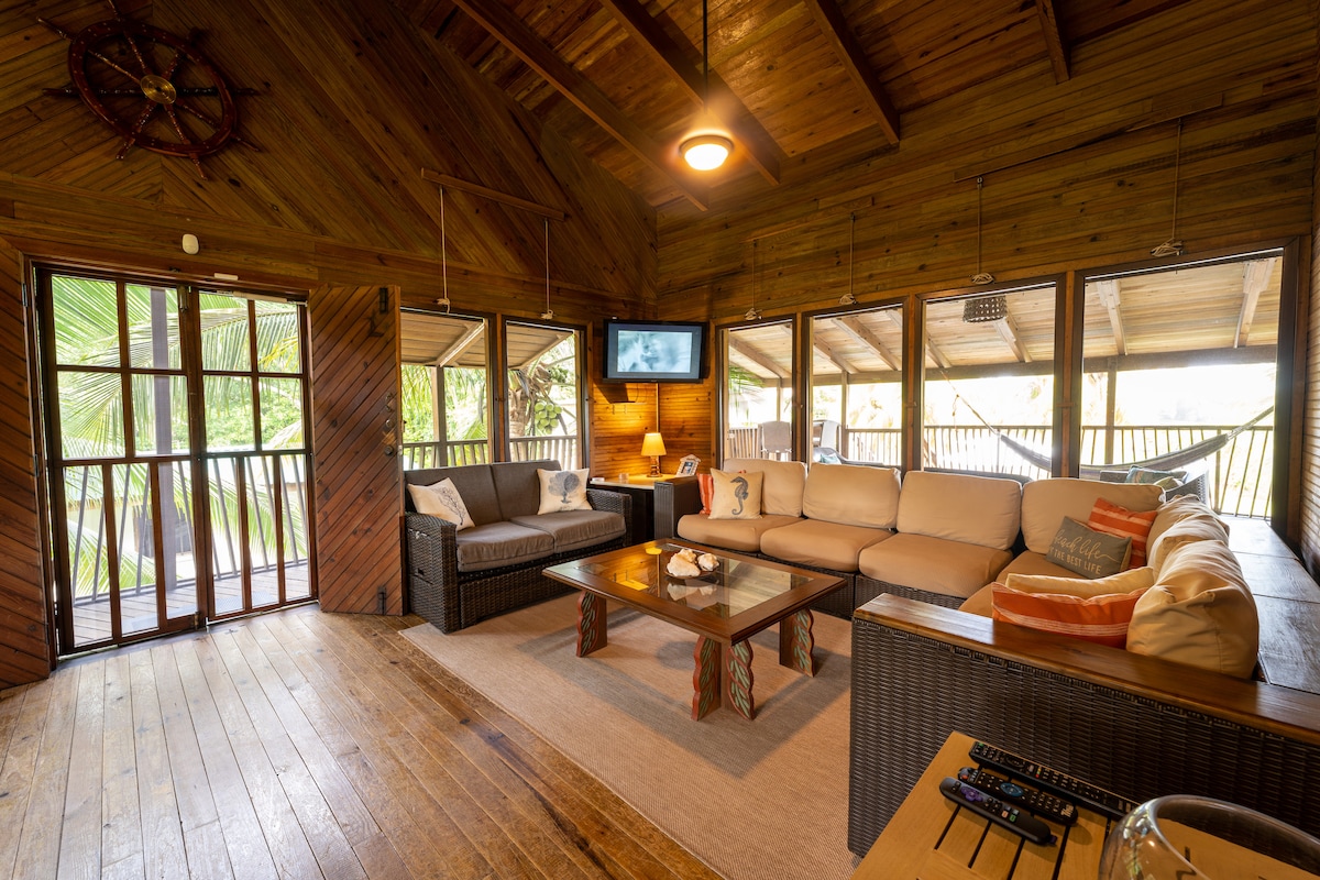 A spacious living area features large windows allowing natural light to fill the room. Comfortable couches surround a wooden coffee table, and a flat-screen TV is mounted on the wall. The wood-paneled walls add warmth to the inviting space.