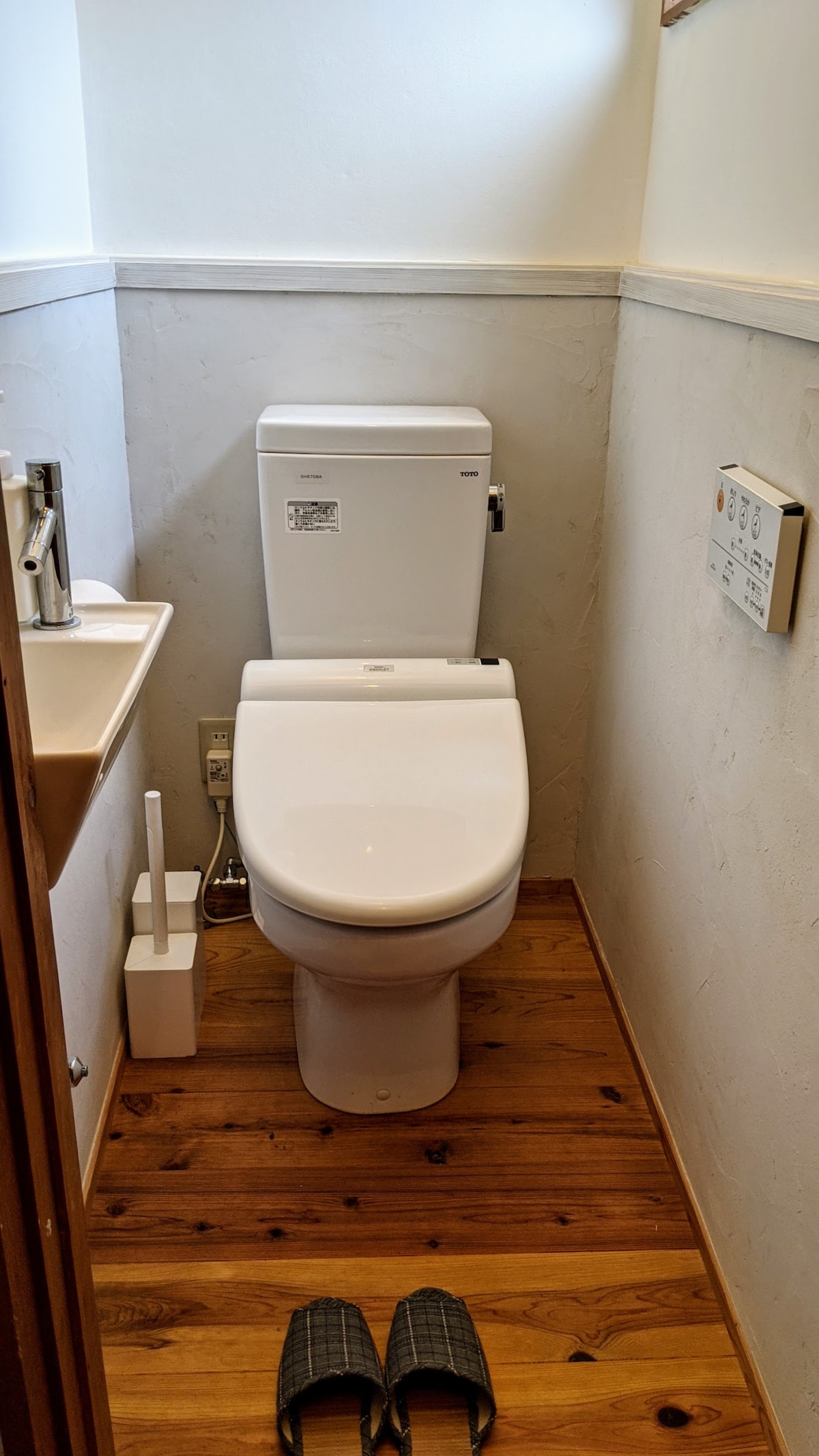 A compact bathroom space features a modern toilet and a small sink with a mirror above it. Wooden flooring complements the neutral wall color, and a pair of slippers is neatly placed on the floor, adding a personal touch to the environment.