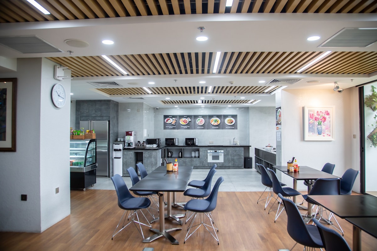A modern dining area features multiple tables and chairs arranged neatly on a hardwood floor. The walls are adorned with artwork, and a kitchen space is visible in the background, equipped with appliances such as a refrigerator and stove. Natural light enters through large windows.