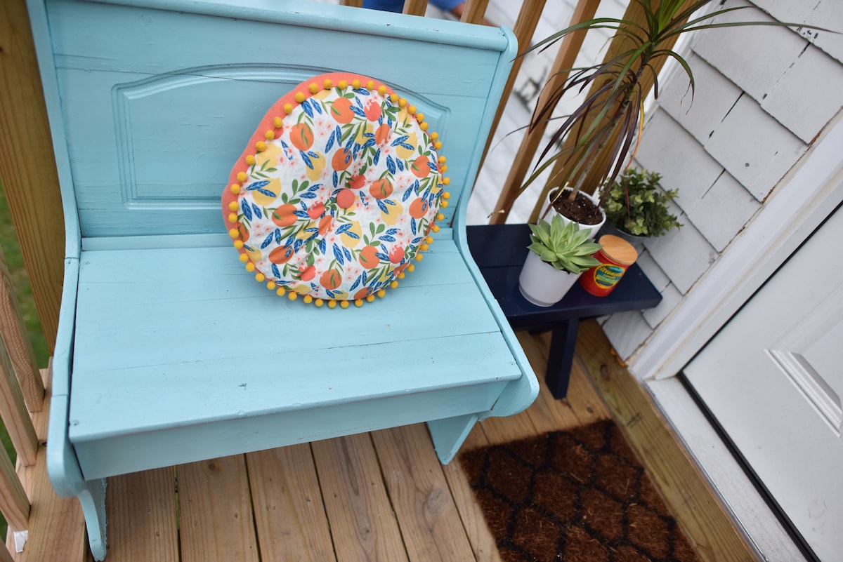 A bright blue bench is situated outside the entrance, adorned with a colorful, floral-patterned cushion. To the side, a small table displays a potted plant and decorative items, enhancing the welcoming atmosphere. A doormat lies in front of the door, complementing the exterior.