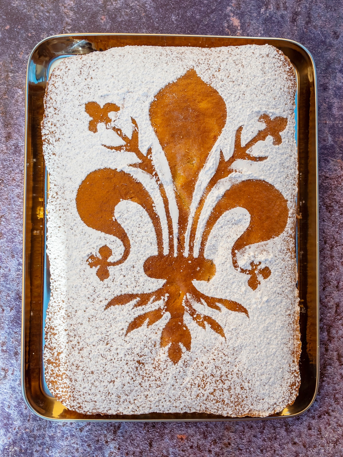 A rectangular tray holds a cake decorated with a powdered sugar design of a traditional fleur-de-lis. The cake's golden-brown surface contrasts with the white sugar, creating an elegant visual presentation.