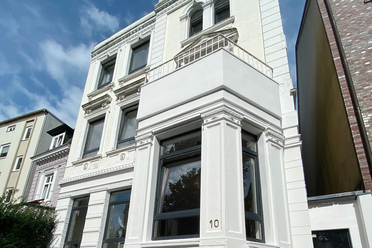 The exterior of a three-story building is presented, characterized by a white facade and decorative architectural features. Large windows allow natural light to enter. The entrance is clearly marked by the number '10'. Surrounding greenery adds to the overall aesthetic.