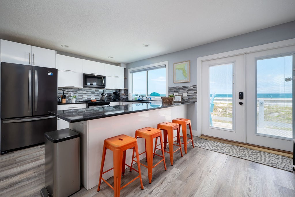 A modern kitchen space features sleek, white cabinetry and dark countertops. Orange bar stools are positioned at the counter. Large windows offer unobstructed views of the ocean, while glass doors provide access to an outdoor area.