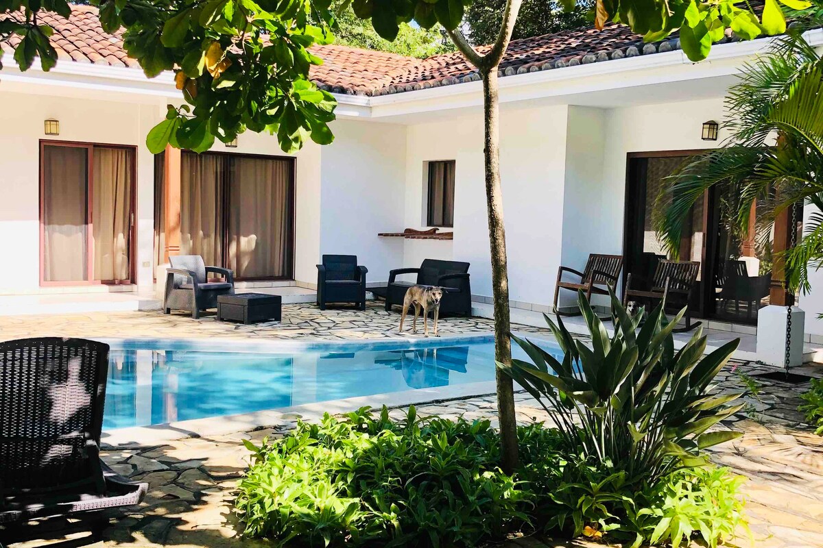 A private pool is surrounded by a stone patio, with comfortable lounge chairs positioned nearby. Lush greenery and tropical plants add a vibrant touch. The home features large sliding glass doors that open onto the pool area, offering easy access to outdoor relaxation.
