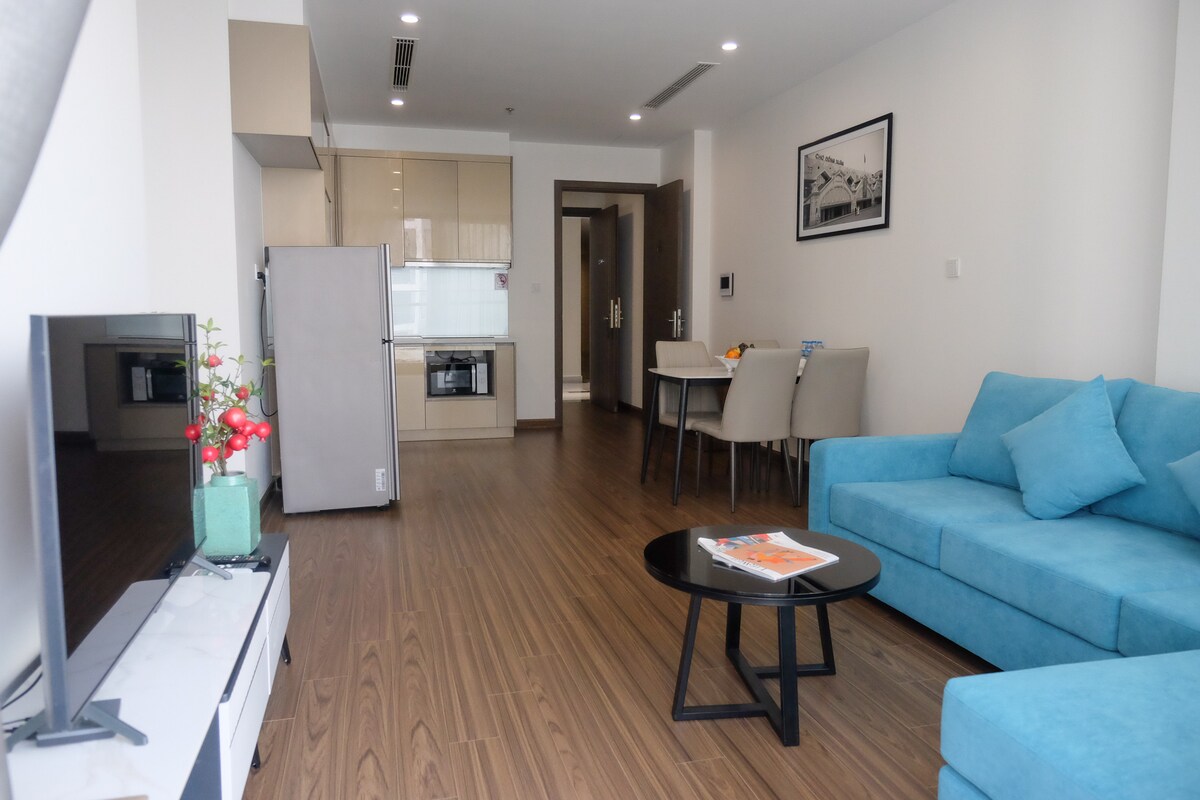 A modern living area is visible, featuring a light blue sofa and a black coffee table. A small dining table with four chairs is situated in the background. The kitchen area includes cabinetry and appliances, while a television is positioned in the living room.