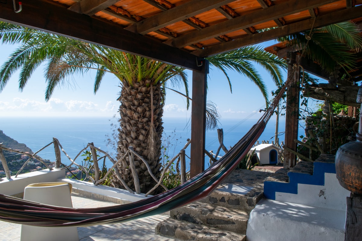 A hammock hangs between wooden posts under a covered terrace, with vibrant green palm leaves swaying nearby. Steps lead down to a lower area, where expansive sea views can be enjoyed in the distance. Sunshine illuminates the serene outdoor space.