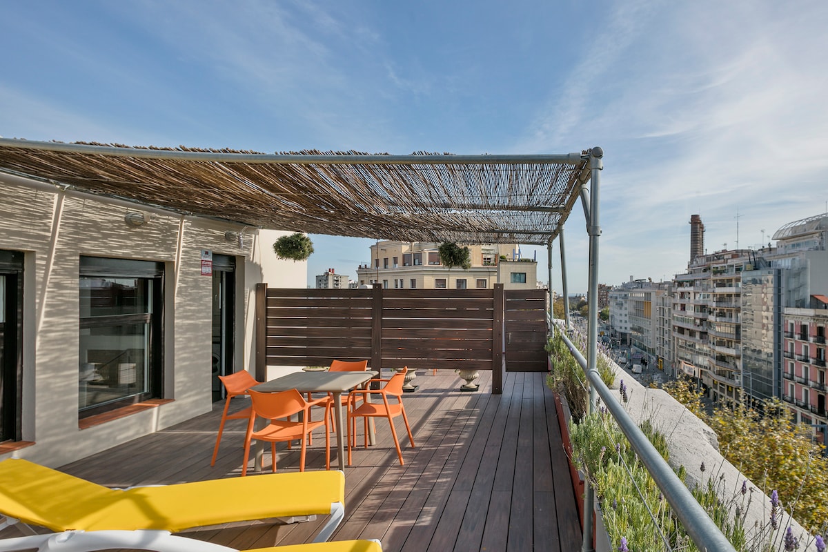 A spacious private terrace features a wooden deck adorned with potted plants. A dining table with orange chairs is positioned under a shade structure, while lounge chairs in the foreground offer a comfortable spot to relax. The view encompasses the city skyline in the background.