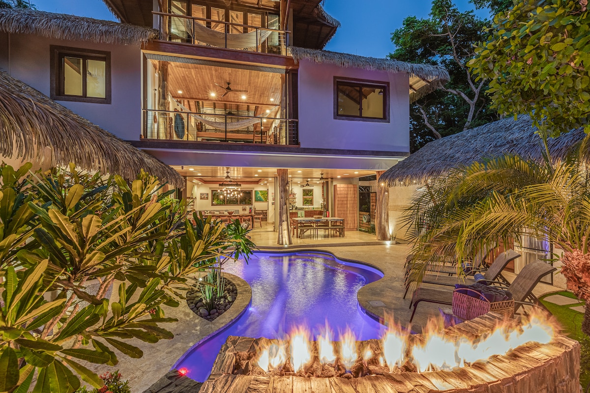 The backyard features a saltwater lagoon pool surrounded by lush tropical foliage. A decorative gas fire feature is positioned above the natural stone edge. The multi-level villa is visible in the background, showcasing an open layout with expansive windows and a thatched roof.