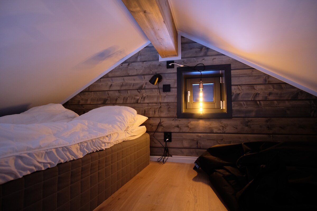 A cozy loft space features a spacious bed covered with white bedding. Warm light emanates from a hanging lamp, highlighting the wooden wall paneling. A dark corner holds a large beanbag. Minimalist decor contributes to a tranquil atmosphere.