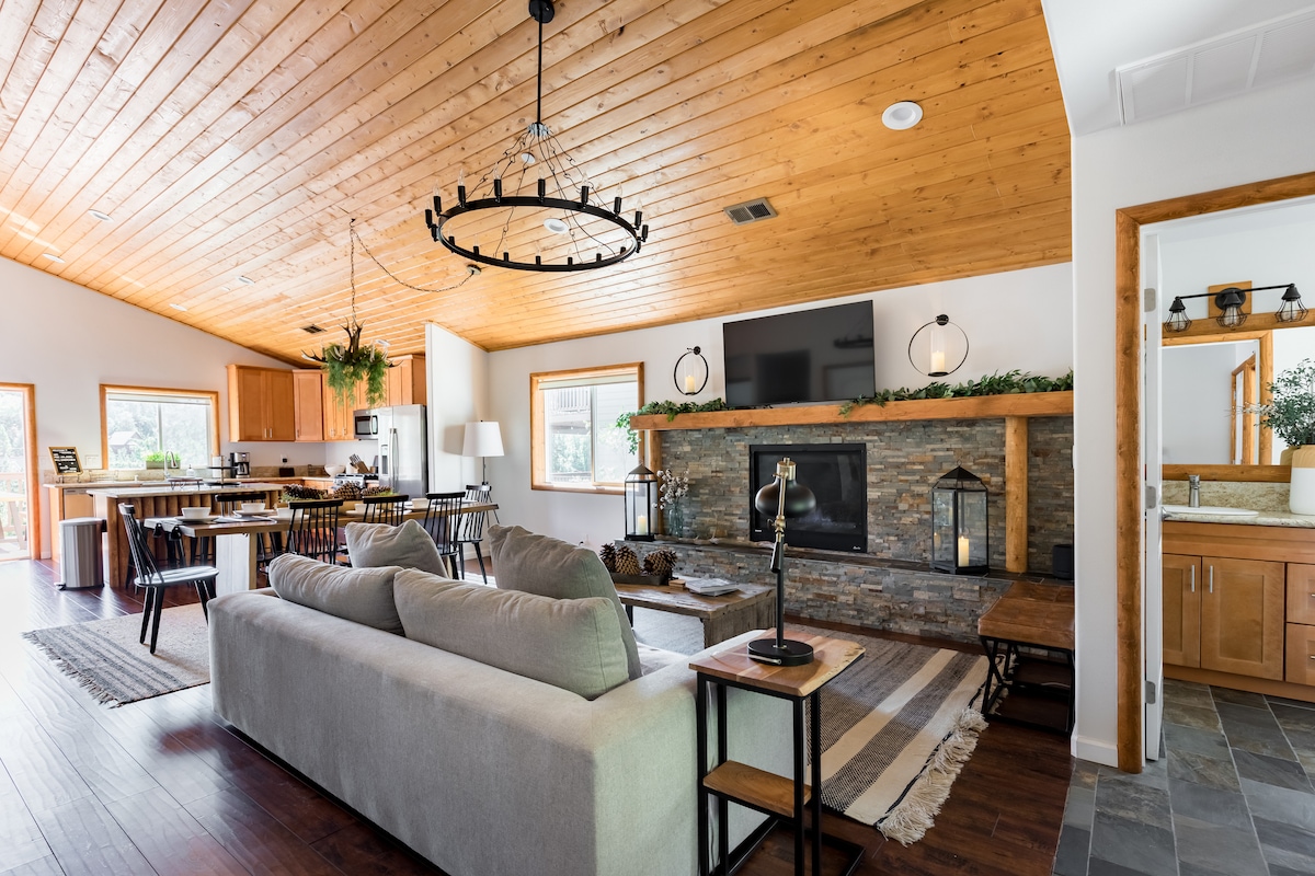 The open-concept living area features vaulted wood ceilings and a stylish gas fireplace with a stone surround. A spacious sectional sofa faces a large flat-screen TV, while a dining table for eight and a kitchen are visible in the background, illuminated by natural light.