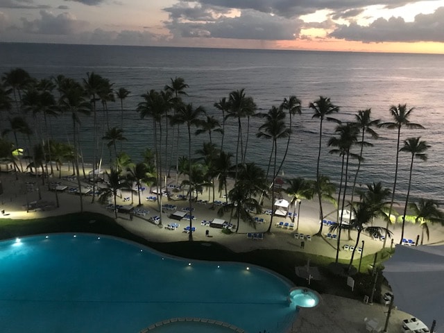 A serene beachfront scene captures a shimmering pool in the foreground, flanked by lounge chairs. Tall palm trees frame the shoreline, and gentle ocean waves reflect the soft hues of a sunset sky.