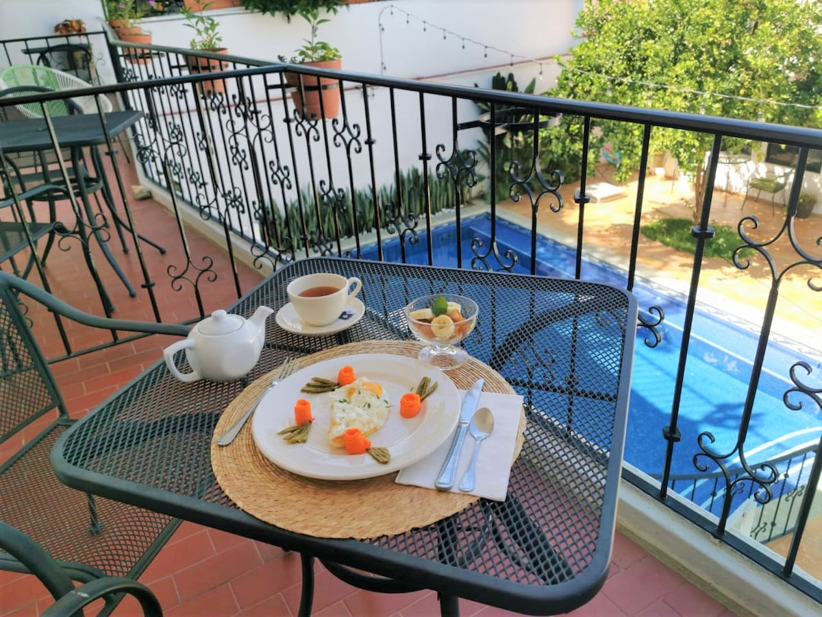 A table set for breakfast is displayed on a balcony, featuring a plate with eggs and colorful vegetables alongside a cup of coffee and fruit. The view includes a blue swimming pool and greenery in the background, framed by decorative railings.