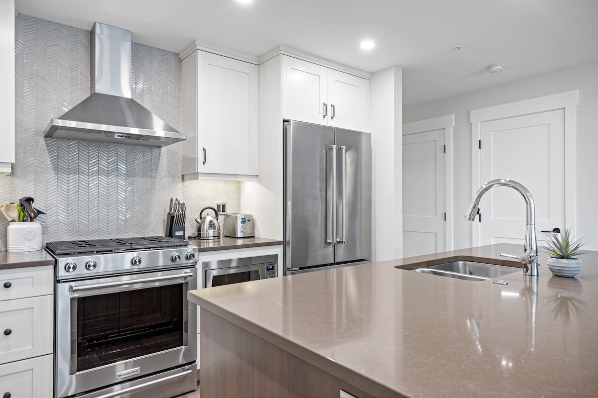 The kitchen features modern stainless steel appliances, including a gas range and a double-door refrigerator. A large island with a sleek countertop provides ample workspace, while light-colored cabinets and a textured backsplash create a bright atmosphere.
