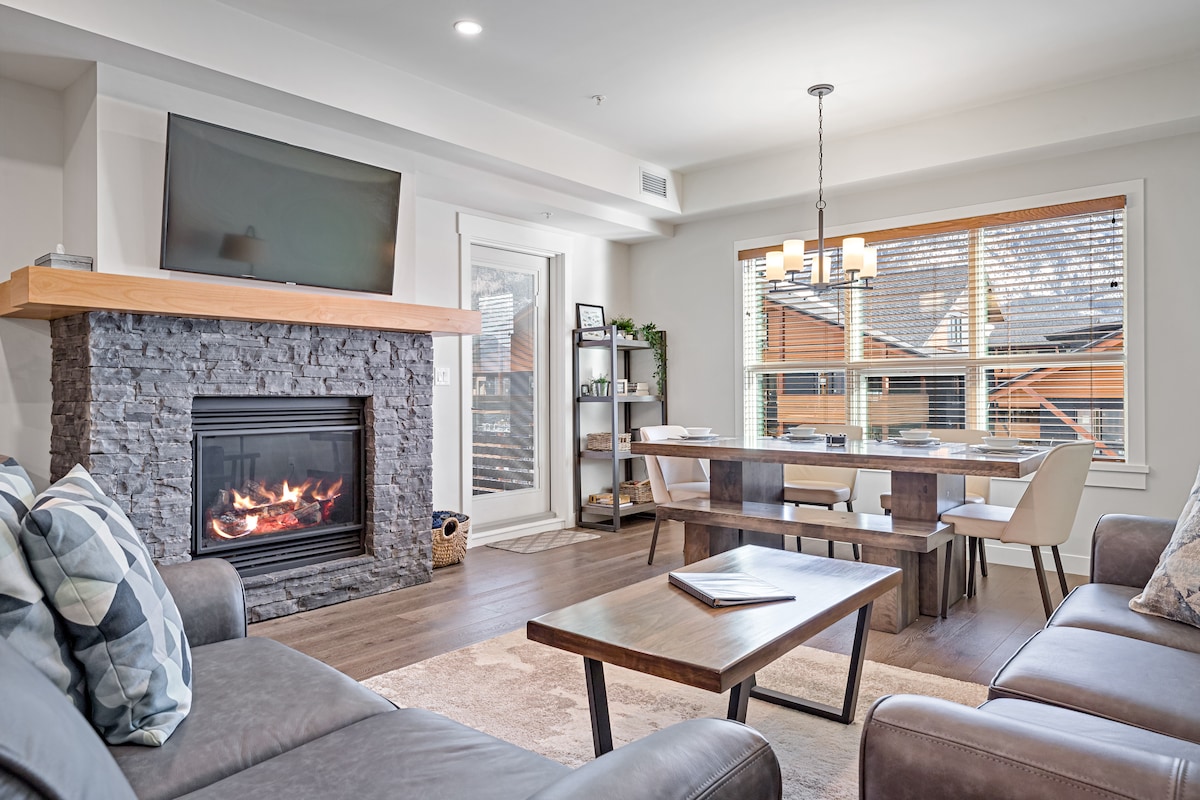 A cozy living area is highlighted by a stone fireplace with a modern television mounted above. Natural light filters through the window, illuminating a dining table with seating for four. Comfortable sofas and a neutral-toned rug enhance the inviting ambiance.
