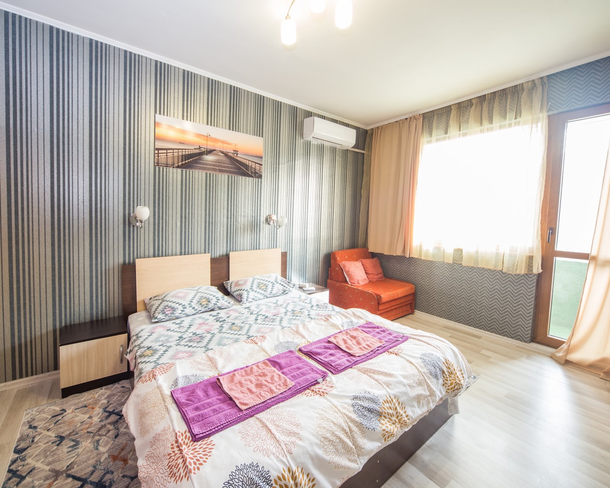 A well-lit bedroom features a spacious bed dressed in a patterned duvet and two throws. An accent wall with soft stripes complements the decor. A small seating area with an orange couch is visible, alongside a large window draped with sheer curtains, allowing natural light to fill the space.