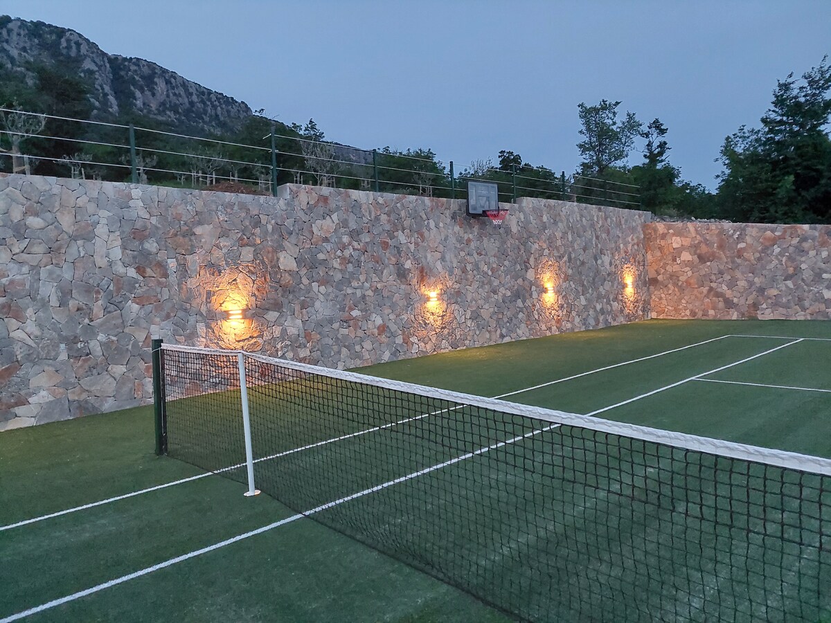 A multi-use sports court is illuminated in the evening, featuring a well-maintained tennis court with a net and clear boundary lines. A stone wall surrounds the court, enhancing the ambiance, while floodlights provide visibility for nighttime play.