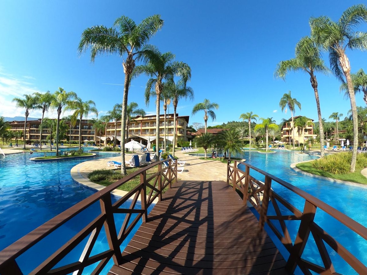 A broad wooden walkway leads through lush palm trees towards a serene pool area. The expansive resort features multiple swimming pools, lounging areas, and landscaped gardens, all under a clear blue sky, creating a tranquil environment for relaxation.