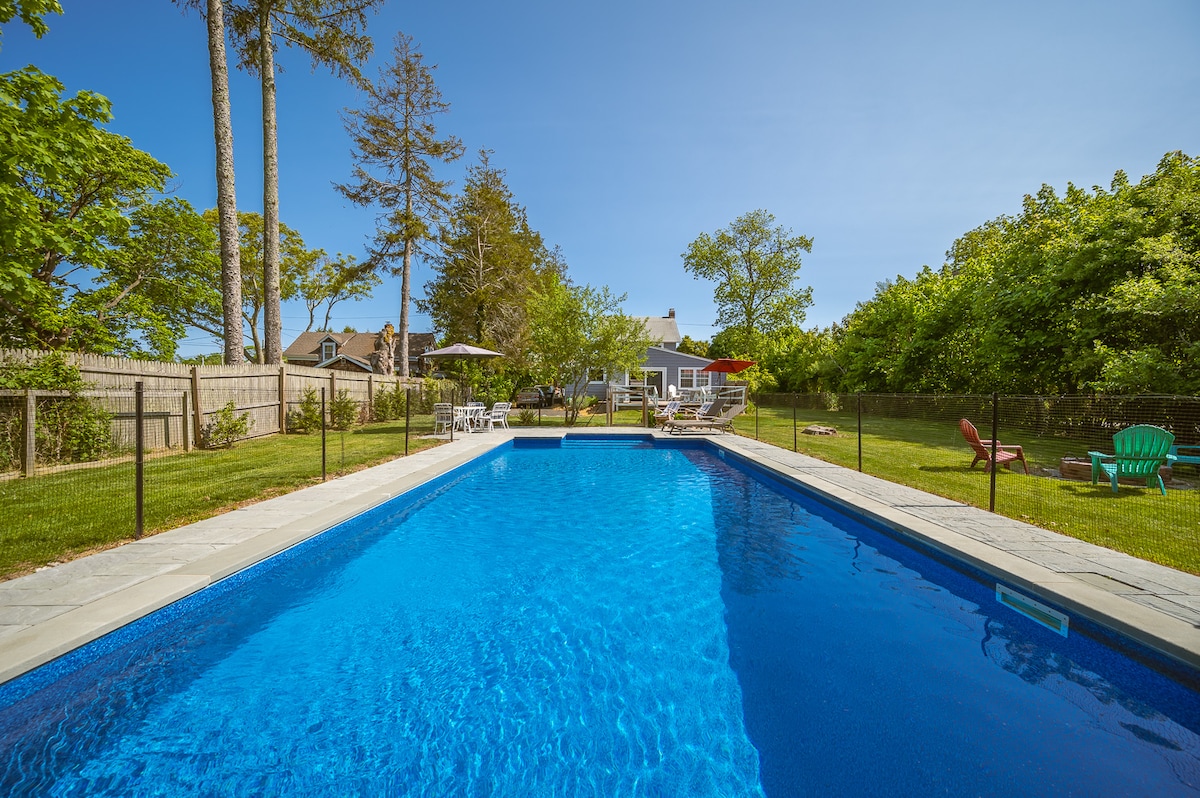 A large in-ground pool is surrounded by a spacious patio, featuring lounge chairs and seating areas. Lush greenery and trees are visible in the background, creating a tranquil outdoor setting. A fence encloses the pool area, ensuring privacy.