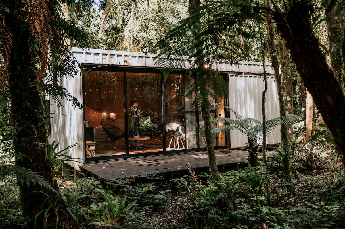 The cabin is surrounded by lush greenery, blending seamlessly into the forest. Large glass panels provide a view of the interior, which features a cozy seating area and warm decorations. Wooden decking extends out, offering a natural connection to the surrounding Mata Atlântica.
