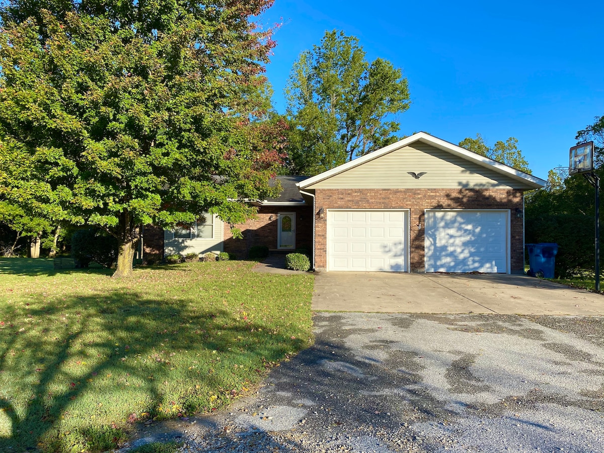 The exterior of a single-story brick home is shown, featuring a two-car garage with double doors. A spacious driveway is visible, accompanied by a well-maintained lawn, mature trees, and a basketball hoop. Clear blue skies create a bright atmosphere.