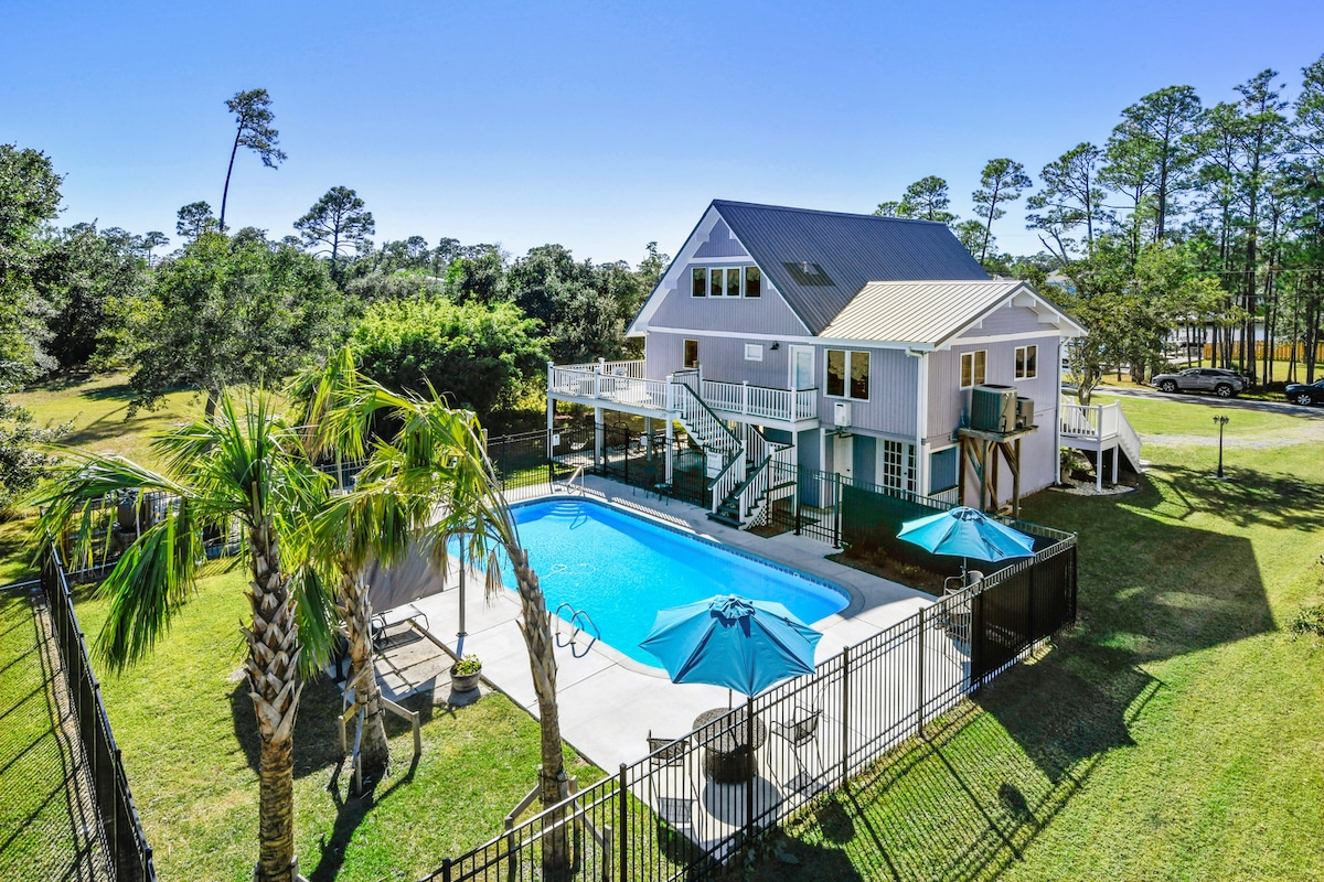 A spacious vacation home is set within a natural landscape, featuring a fenced heated pool. Umbrellas provide shade around the pool area, while palm trees frame the exterior. The home's multiple decks overlook the surrounding greenery and can be accessed via outdoor stairs.