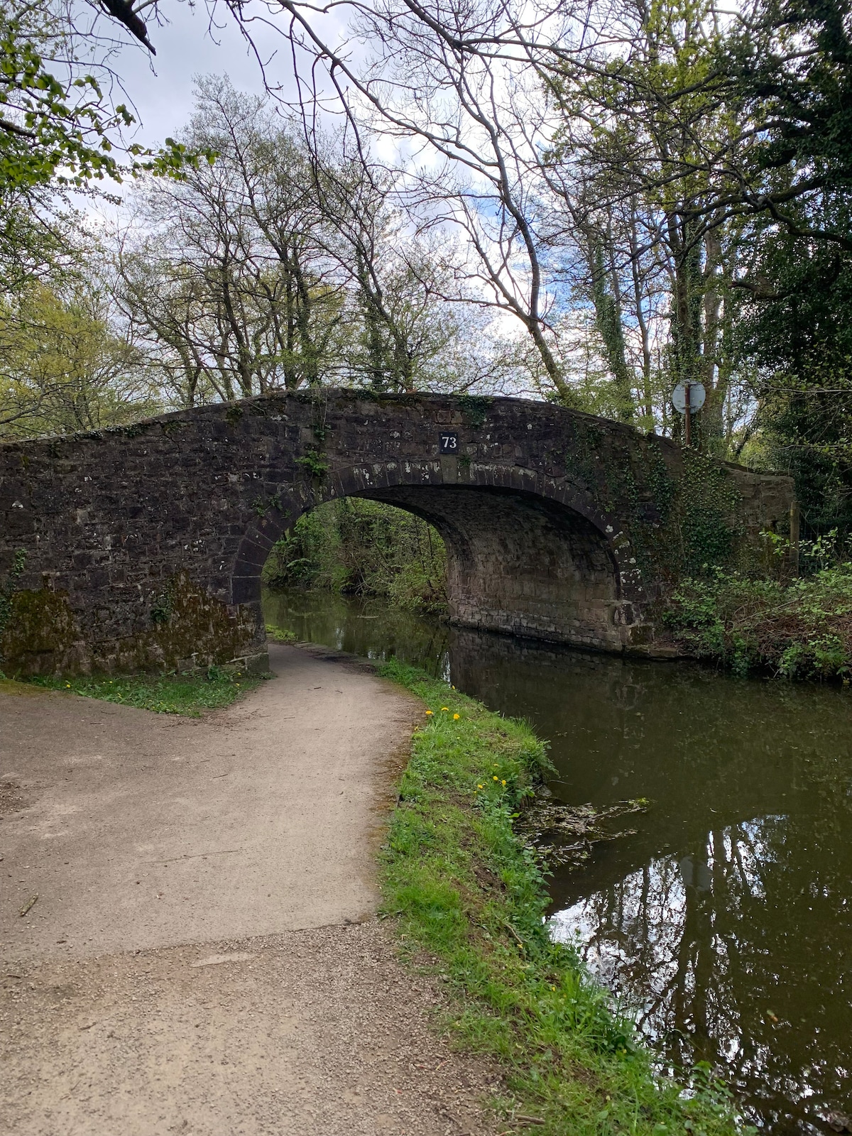 A stone arch bridge spans a calm canal, surrounded by lush greenery. A path runs alongside the water, inviting leisurely walks. Trees with budding leaves provide a gentle canopy above, while the water reflects the serene landscape. Road signage is visible near the bridge.