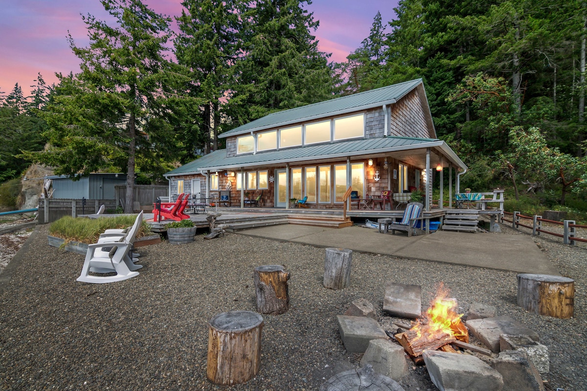 The cabin-style home is surrounded by trees and faces a sandy area, featuring large windows that provide views of the outdoors. A fire pit is visible in the foreground, with log seating arranged around it, complemented by chairs in vibrant colors. A serene atmosphere is created by the natural setting.