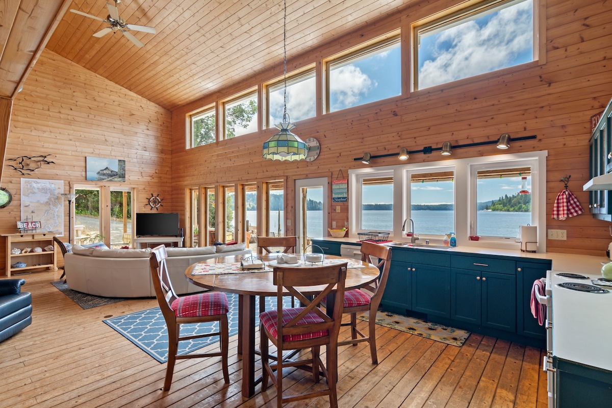 An open floor plan cabin features large windows allowing natural light to fill the space. A round dining table sits center, surrounded by wooden chairs with colorful cushions. The kitchen area is equipped with dark cabinetry, offering a view of the waterfront through expansive glass panes.