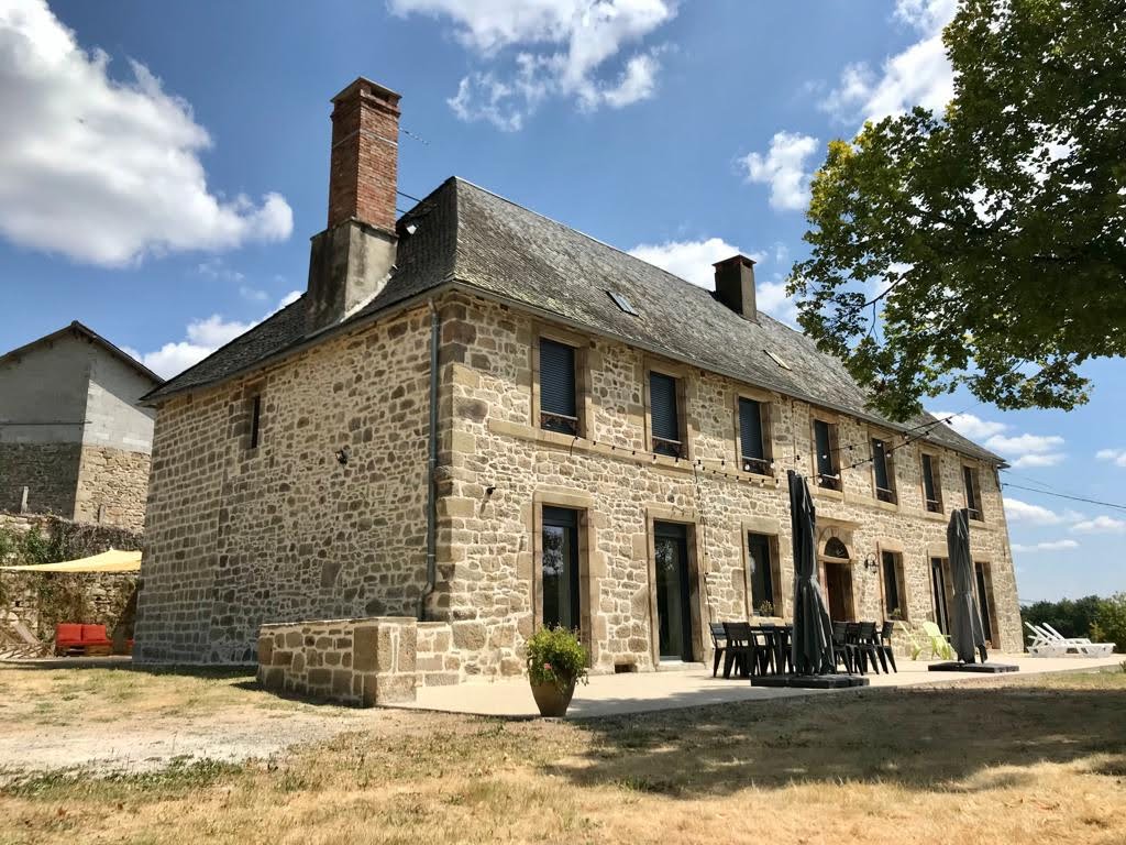 A spacious stone house is depicted against a bright sky, showcasing its traditional architecture. A large terrace with dining furniture is visible in front, providing access to the surrounding nature. Umbrellas stand ready for shade, enhancing outdoor leisure opportunities.