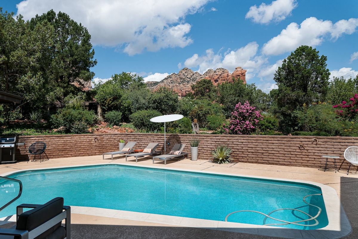 A serene backyard pool area is showcased, featuring a blue pool bordered by light-colored stone. Lounge chairs and an umbrella are positioned nearby, with lush landscaping and vibrant flowers in the background, all framed by the stunning Red Rock mountain views.