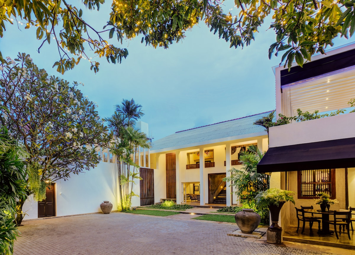 A serene outdoor area is framed by lush greenery and illuminated by soft lighting. The modern architecture features a blend of light-colored walls and a terracotta roof, while a stone pathway leads to a spacious entrance, creating an inviting atmosphere.