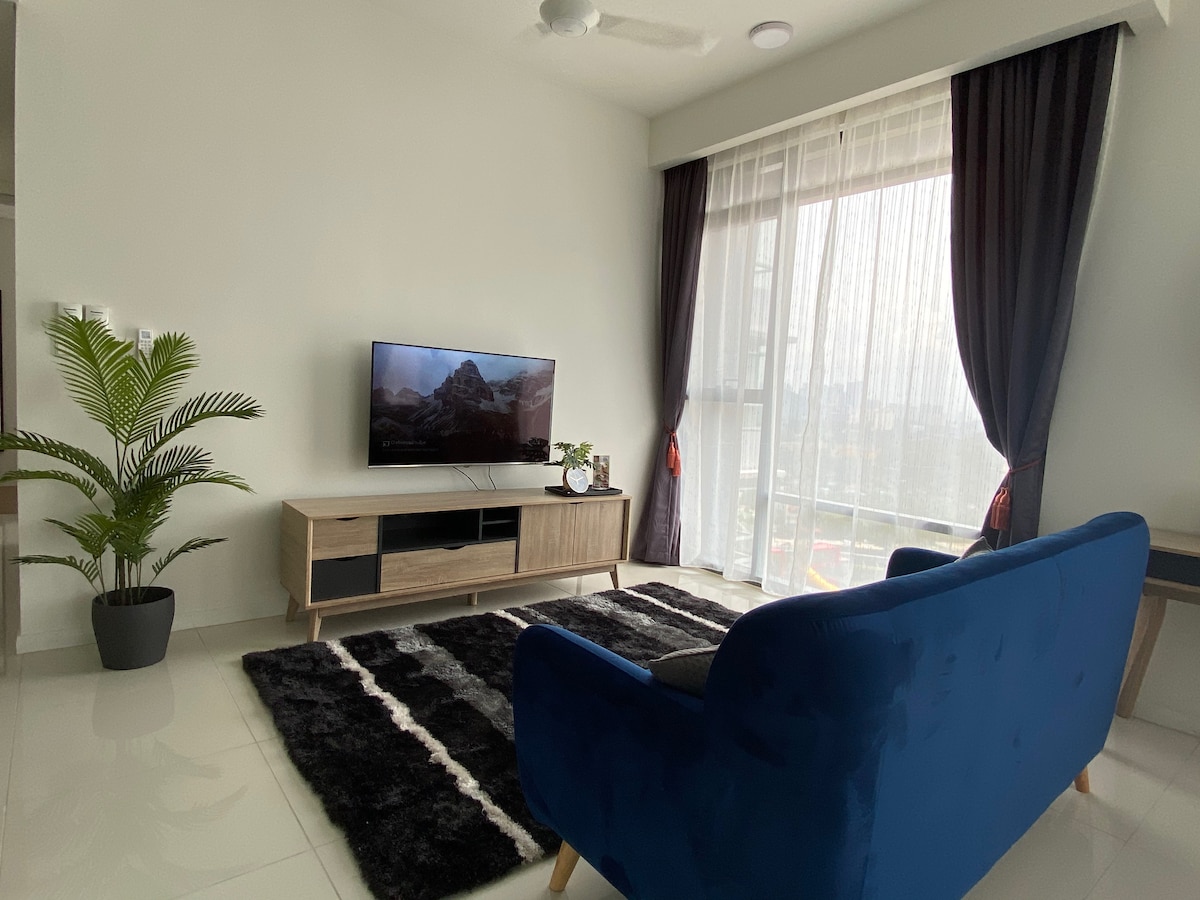 A living area features a blue three-seater sofa positioned across from a wall-mounted Smart TV. A black and white area rug covers the floor, while a potted plant adds greenery. Natural light streams in through sheer curtains, creating a bright and open atmosphere.