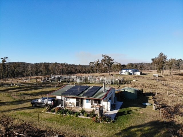 An aerial view captures a rural property featuring a cottage with a solar panel roof, surrounded by open fields and grazing area. Nearby structures include a water tank and fencing, with trees lining the horizon under a clear blue sky.