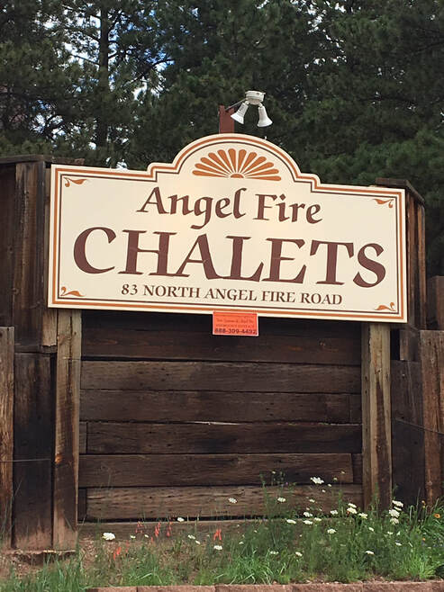 A wooden sign displaying 'Angel Fire Chalets' is prominently featured, set against a backdrop of greenery. The sign is bordered with an elegant design, providing clear visibility of the location. The ground is adorned with wildflowers, adding a touch of nature to the scene.