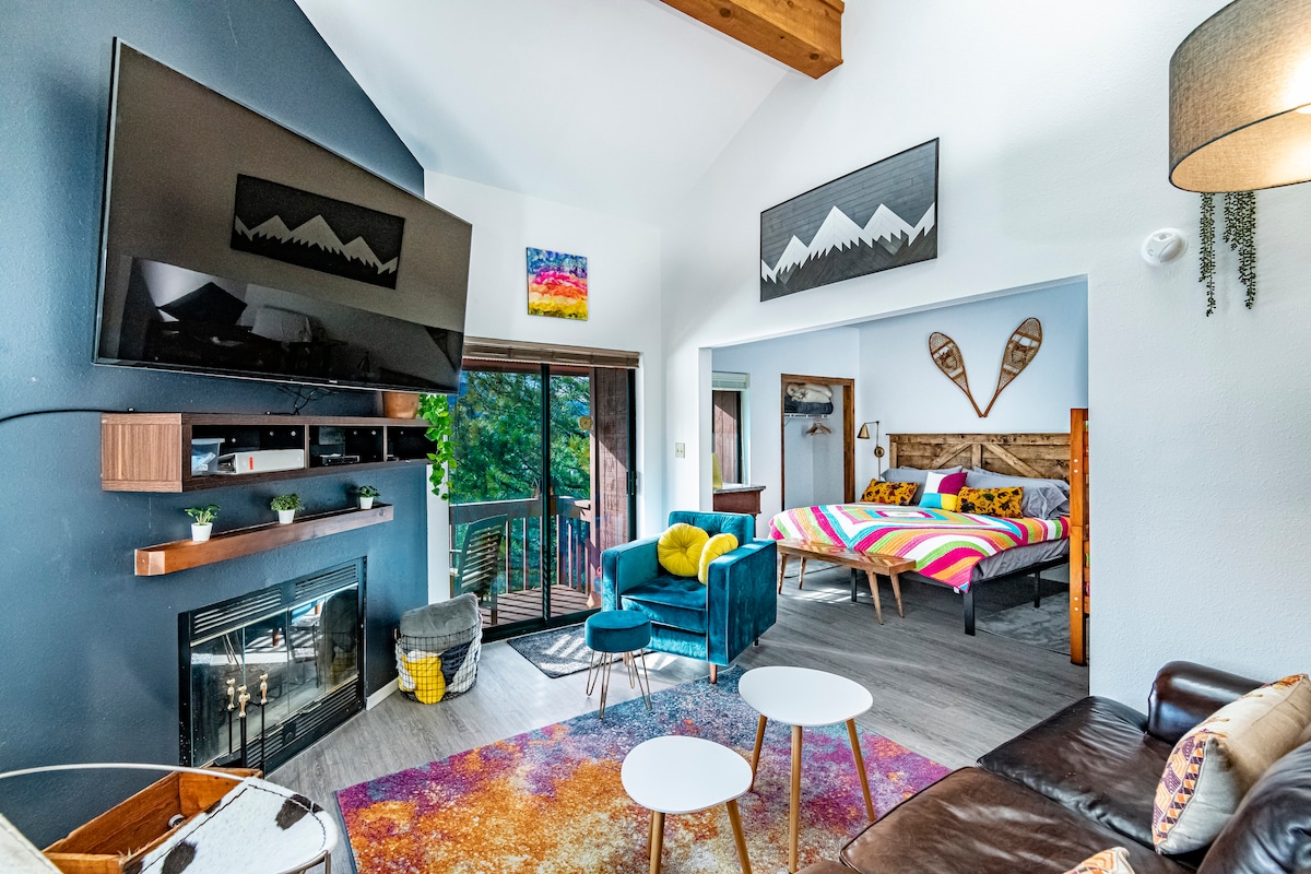 The living area features a wood-burning fireplace below a large TV mounted on the wall. A sliding glass door opens to the balcony, while a colorful rug enhances the space. A cozy seating area includes two chairs and a coffee table, with a glimpse of the adjacent bedroom.