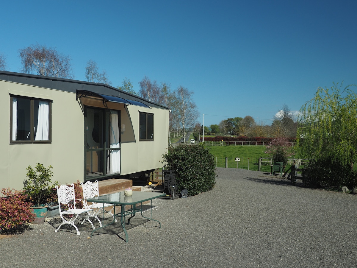 A vintage mobile home is situated in a serene outdoor setting, featuring a small porch with steps. A simple seating area with white chairs and a glass table is visible. Lush greenery and open fields are seen in the background under a clear blue sky.