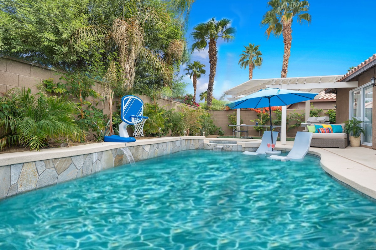 A refreshing pool is surrounded by lush greenery and tropical palm trees, featuring an in-pool lounger for relaxation. A blue umbrella provides shade over outdoor seating. A basketball hoop is positioned at the edge of the pool, enhancing recreational opportunities.