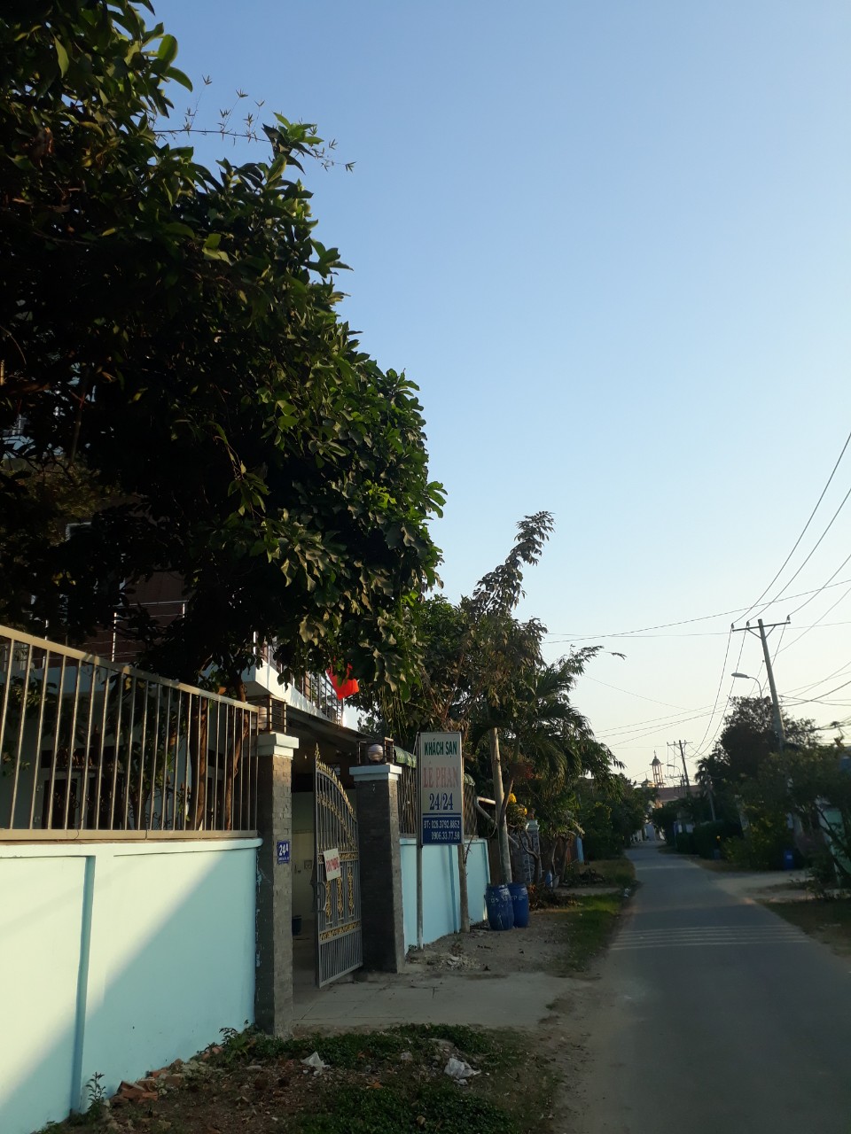 A quiet street is shown, featuring a well-kept entrance with a gated fence. Lush greenery is present along both sides of the road, and clear blue skies are visible above. The surrounding area includes a mix of residential structures and open spaces.