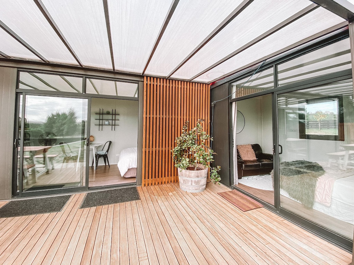 A spacious outdoor deck is framed by large glass sliding doors, providing access to two adjoining pods. A potted plant adds a touch of greenery, while the wooden floor enhances the natural ambiance. The structure features a modern design with a shaded roof overhead.