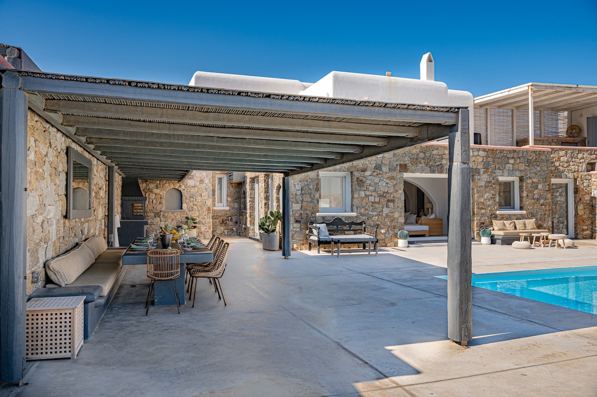 An outdoor dining area is presented under a covered terrace, featuring a long table with comfortable seating. Natural stone walls create a rustic ambiance, while the private pool is visible beyond. Sunlight highlights the serene space, perfect for al fresco meals and gatherings.