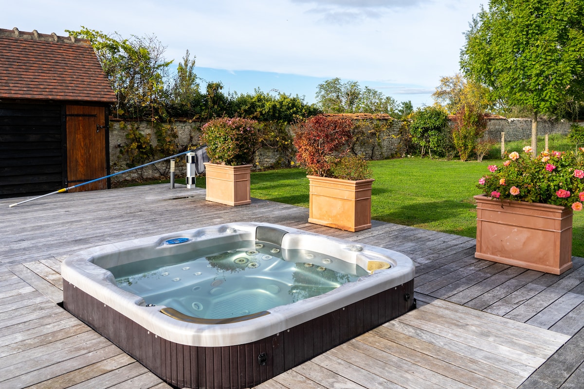 A jacuzzi sits on a wooden deck, surrounded by green grass and flower planters filled with vibrant blooms. Nearby, a small shed is visible in the background, with trees and hedges outlining the property.