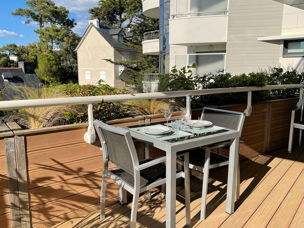 An outdoor terrace is presented with a dining table set for two, featuring clear glass plates and wine glasses. Surrounding greenery adds a natural touch, while modern buildings are visible in the background under a clear blue sky.