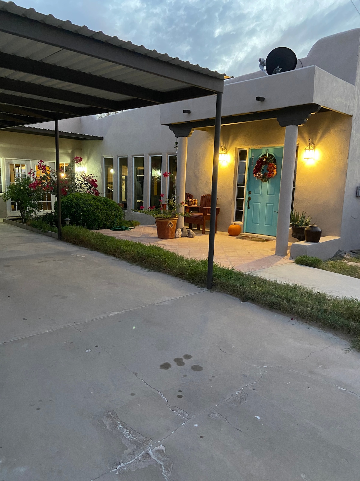 A welcoming entrance is framed by vibrant flowers and potted plants. The light blue door stands out against the neutral facade, illuminated by warm outdoor lights. A shaded porch provides seating, while a pathway leads into the home, offering a charming glimpse of the outdoor space.