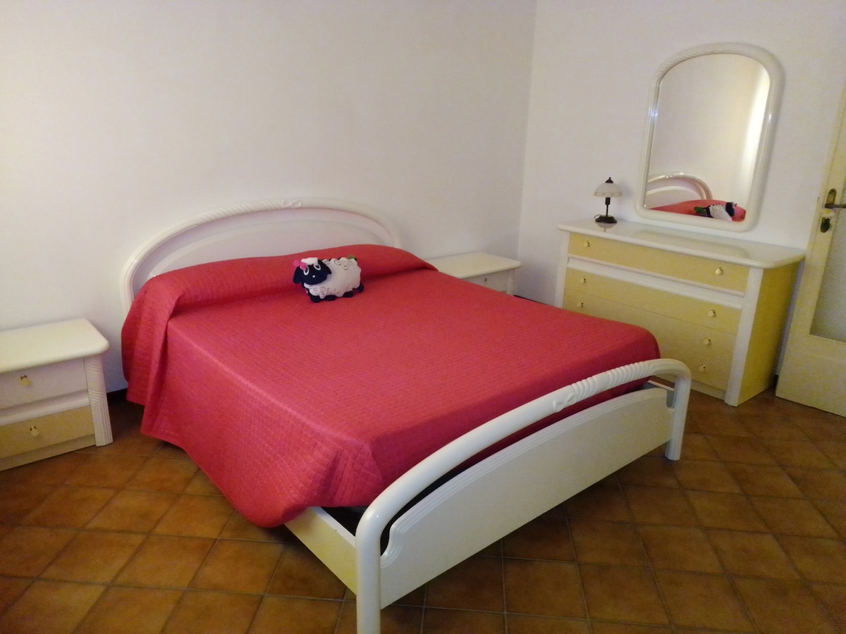 A spacious bedroom features a large red bedspread covering a generous bed, with a decorative plush sheep sitting atop. Two nightstands flanking the bed and a dresser with a mirror are visible, all arranged in a simple, clean layout on tiled flooring.
