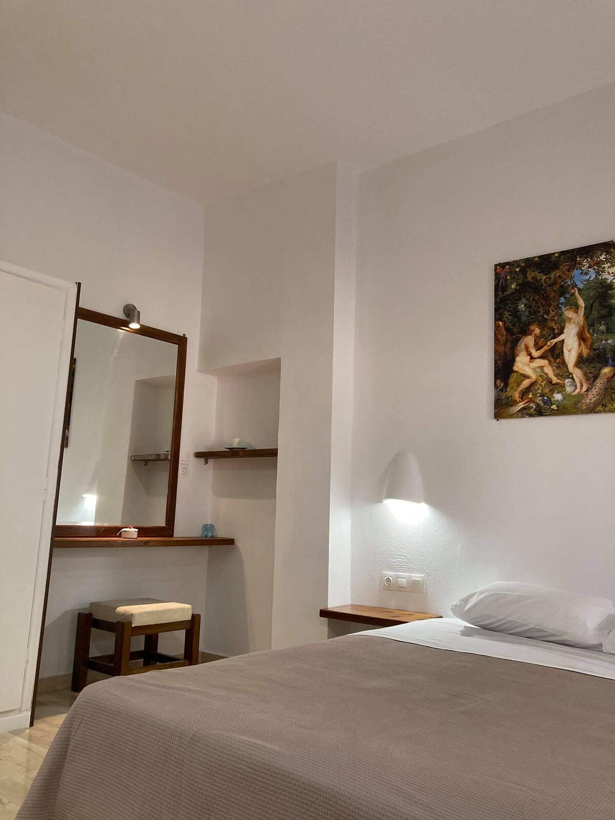 The room features a spacious bed with neutral bedding, complemented by a wooden seating area and a framed artwork on the wall. A mirror above the dressing table adds functionality, while soft lighting highlights the clean lines of the interior.