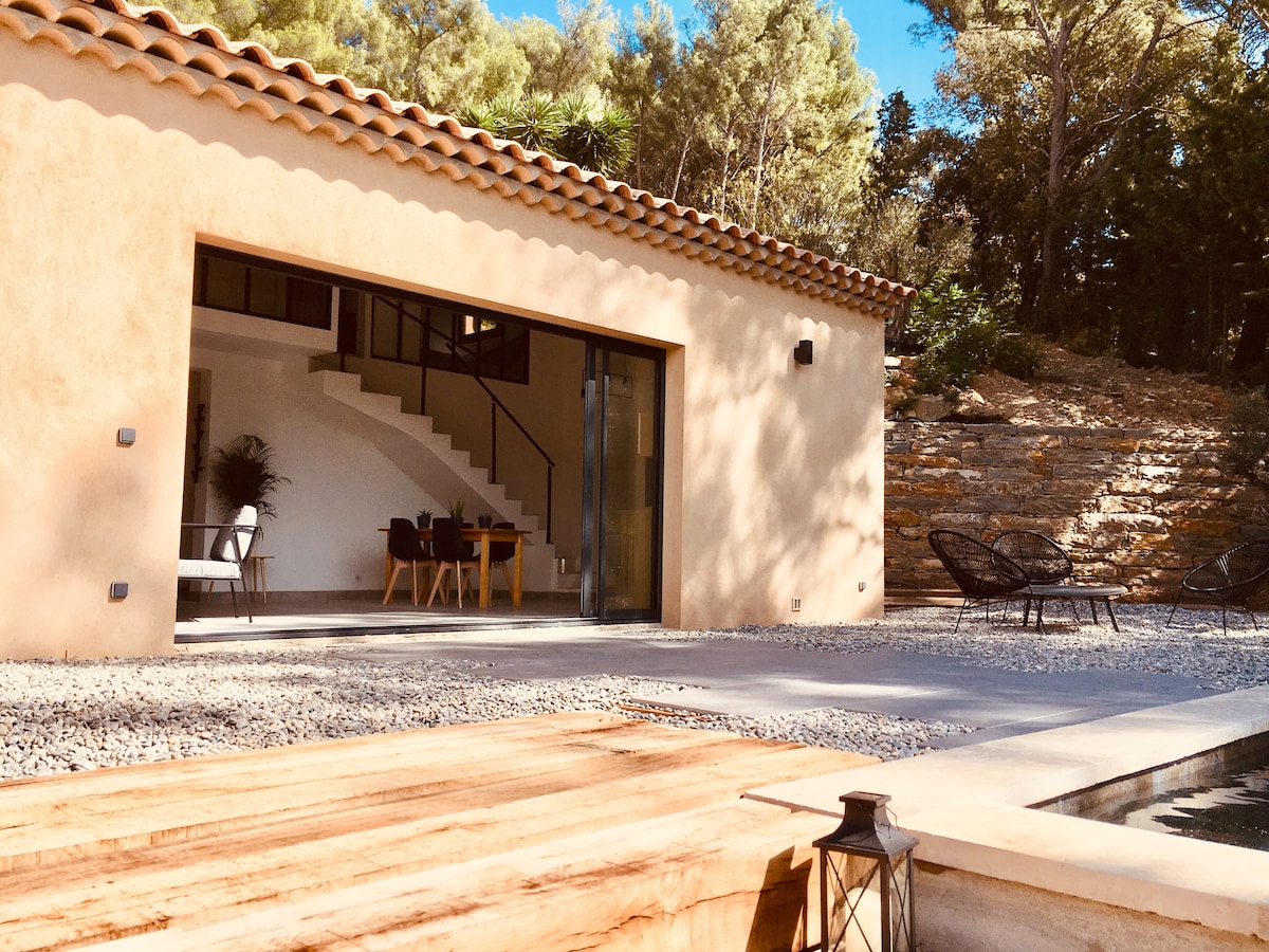 A modern villa features a spacious outdoor terrace with a wooden deck that leads to a private swimming pool. Large glass doors open to the interior, showcasing a bright living area and a stylish staircase. Lush greenery surrounds the property, offering a natural ambiance.