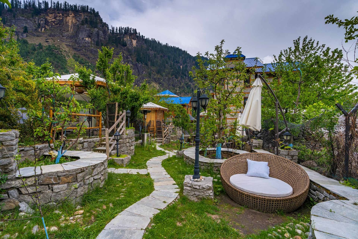 A landscaped garden area is depicted with winding stone pathways surrounded by greenery. A circular seating arrangement features a plush white cushion, while a stone fire pit is visible nearby. The backdrop showcases mountain views and structures with blue roofs.