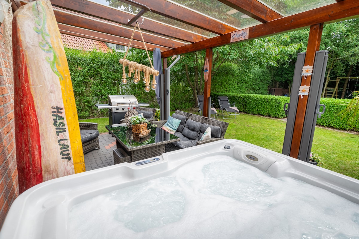 A spacious covered terrace features a large whirlpool, inviting for relaxation. Nearby, a grill is present alongside comfortable lounge furniture surrounded by greenery, creating a serene outdoor environment. Decorative elements enhance the space, complementing the overall inviting atmosphere.