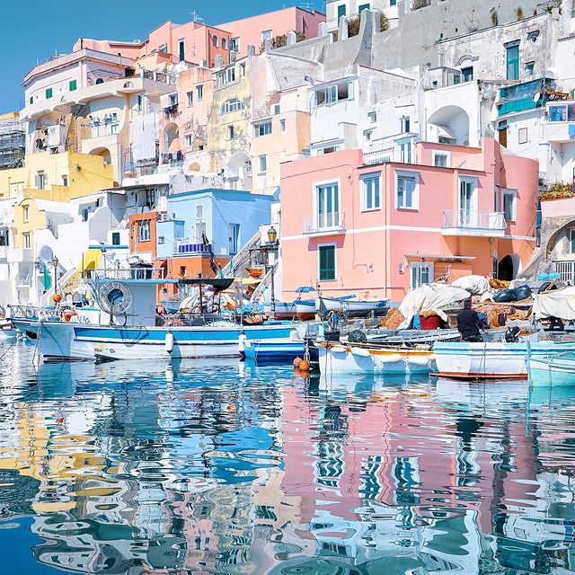 Colorful buildings line the waterfront, reflected in the calm, blue waters. Boats are moored along the dock, surrounded by fishing nets and equipment. The scene captures the vibrant charm of the coastal landscape.