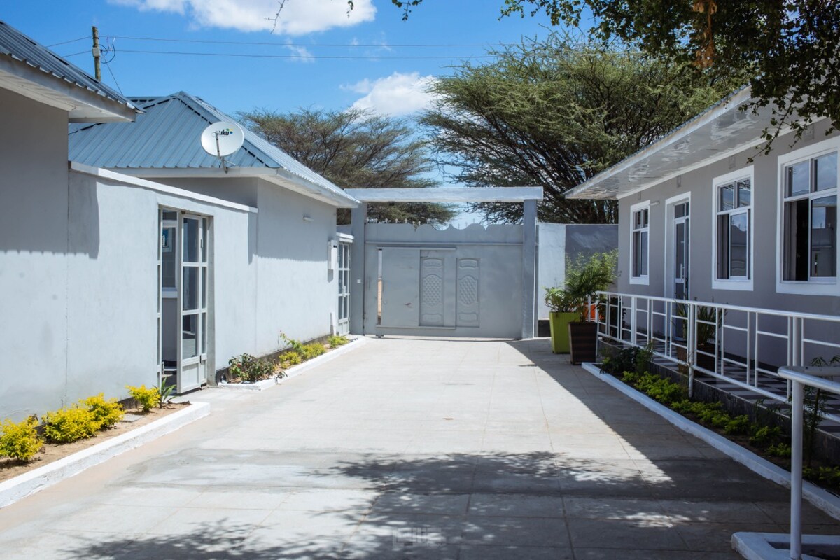 A well-maintained pathway leads through a spacious outdoor area, flanked by two modern buildings with large windows. Lush greenery and freshly planted flowers enhance the environment. A secure entrance gate stands at the far end, providing a sense of privacy and safety.